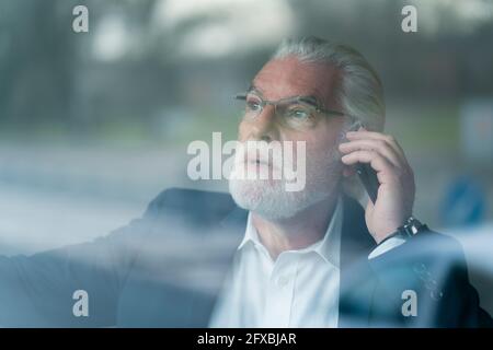 Surprised senior businessman talking on smart phone at office seen through glass Stock Photo