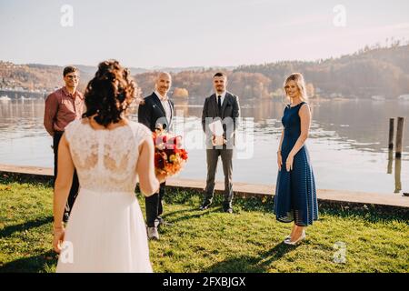 Groom standing with wedding guests looking at bride in front of lake on wedding ceremony Stock Photo
