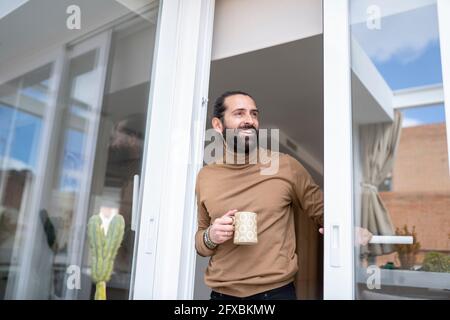 Smiling man with mug looking away while standing at doorway Stock Photo