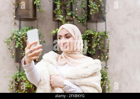 Young woman taking selfie through mobile phone in front of wall Stock Photo