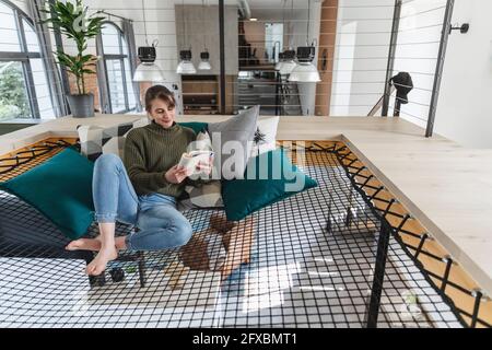 Woman reading book while sitting on hammock in modern home Stock Photo