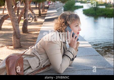 Pretty and elegant middle-aged woman talking on the mobile phone in a park leaning against the wall on the bank of a river in the city Stock Photo