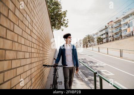 Smiling handsome man looking away while walking with bicycle on footpath Stock Photo