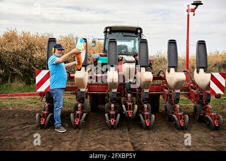 Male farmer wearing ear protectors while pouring corn in containers of tractor at agricultural field Stock Photo