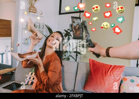 Smiling female influencer showing high heels while filming by friend through mobile phone by social icons at home Stock Photo