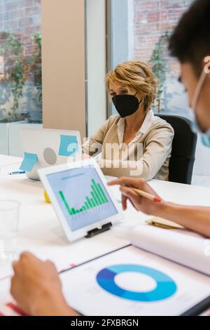 Businessman working on financial data over digital tablet while female coworker using laptop at desk in office during COVID-19 Stock Photo