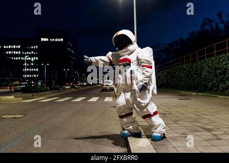 Female astronaut in space suit hailing ride while standing on footpath near road at night Stock Photo