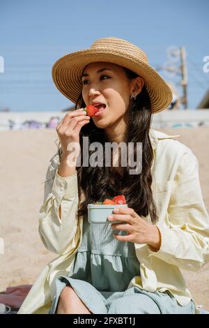 Beautiful woman wearing hat eating strawberries while sitting at beach on sunny day Stock Photo