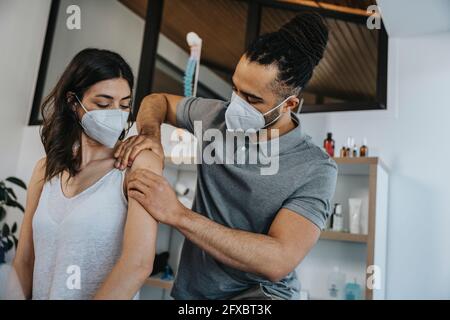 Male physiotherapist working on shoulder of female patient in practice during COVID-19 Stock Photo