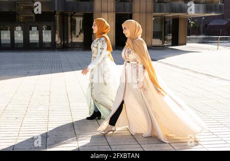 Fashionable friends wearing hijabs smiling while walking on footpath Stock Photo