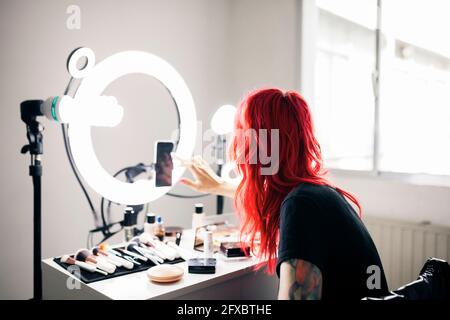 Female artist with dyed red hair using smart phone while vlogging at studio Stock Photo