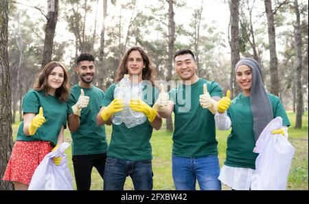 Young volunteer team gesturing thumbs up while holding plastic bottle in forest Stock Photo