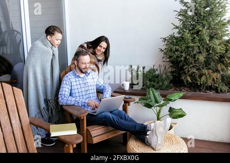 Son and mother looking at father using laptop on balcony Stock Photo