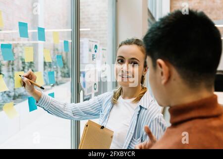 Smiling female and male entrepreneurs discussing over adhesive notes on glass wall at coworking office Stock Photo