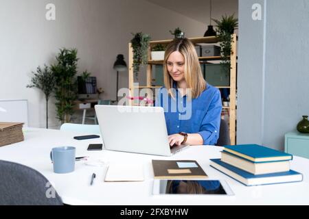 Smiling businesswoman with books on desk using laptop at office Stock Photo
