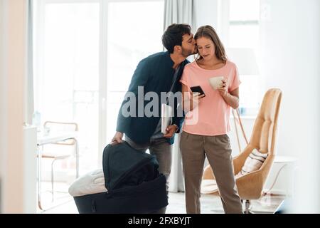 Male entrepreneur carrying baby carrier kissing woman using smart phone in living room Stock Photo