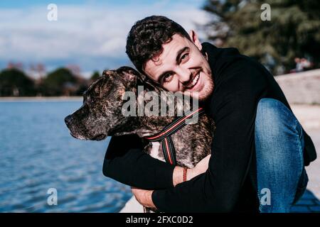 Smiling young man hugging dog while crouching by lake Stock Photo