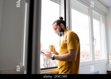 Handsome man using smart phone while standing near window at home Stock Photo