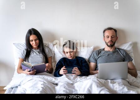 Woman reading book while father and son using wireless technologies in bed at home Stock Photo