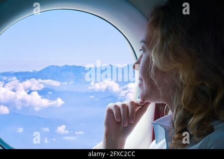 young woman flies in a plane looking through the porthole at the mountains below Stock Photo