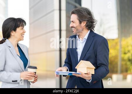 Smiling male real estate agent with house model discussing with female customer holding disposable cup in park Stock Photo