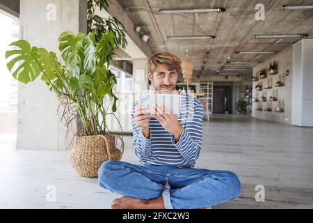 Man holding digital tablet while sitting on floor by plant at loft Stock Photo