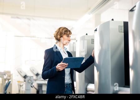 Female manager with digital tablet checking machine while standing in factory Stock Photo