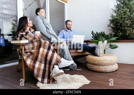 Mother and son wrapped in blanket while sitting with father using laptop on balcony Stock Photo