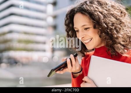 Smiling young woman talking on smart phone while holding laptop during sunny day Stock Photo
