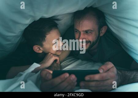 Father and son looking at each other while using smart phone under blanket at home Stock Photo