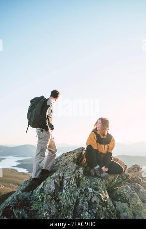 Young female hiker sitting by man standing on top of mountain Stock Photo