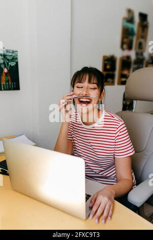 Cheerful woman playing with pen while sitting with laptop at home office Stock Photo