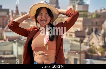 Smiling woman in cap standing with hands behind head at roof terrace Stock Photo