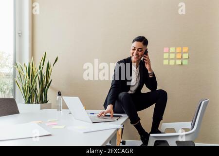 Female entrepreneur talking on mobile phone while using laptop at office Stock Photo