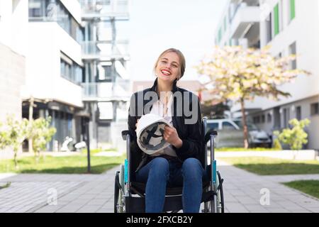 Disabled woman sitting in wheelchair in residential area holding hard hat Stock Photo