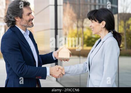 Smiling businesswoman doing handshake with real estate developer holding house model Stock Photo