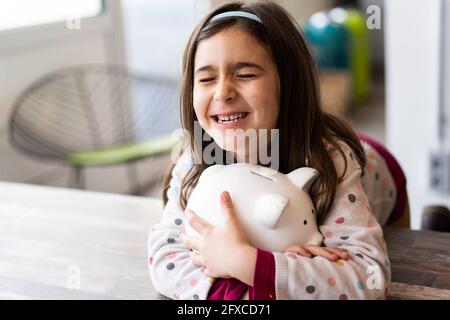 Cheerful girl hugging piggy bank on table Stock Photo