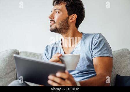 Young man sitting with coffee cup and tablet while looking away Stock Photo