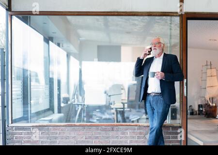 Businessman with coffee cup talking on smart phone while leaning on glass wall at office Stock Photo