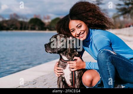 Smiling curly hair woman crouching by dog on sunny day Stock Photo