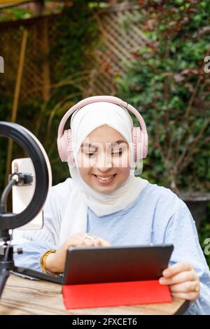 Smiling female influencer wearing headphones using tablet while vlogging at cafe Stock Photo