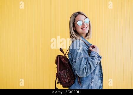 Smiling blond woman with backpack wearing sunglasses by yellow wall Stock Photo