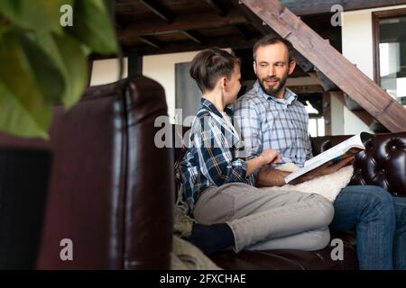 Father holding book while looking at son sitting on sofa at home Stock Photo