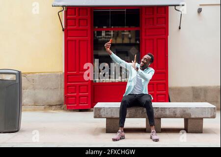 Young man waving during video call while sitting on bench outside store Stock Photo