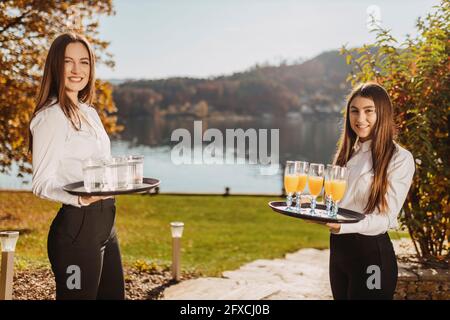 Waitresses standing with tray in garden by lake during sunny day Stock Photo