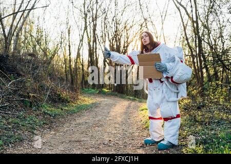 Female astronaut in space suit gesturing while holding cardboard on dirt road Stock Photo