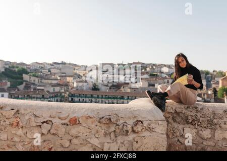 Woman checking map while sitting on retaining wall during sunny day Stock Photo