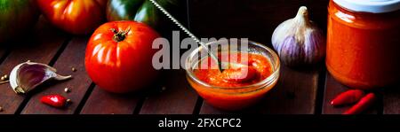 Homemade hot tomato sauce adjika in jars - Tomatoes, chilli pepper, garlic, herbs on wooden table. Stock Photo