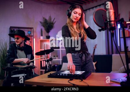 Adorable young woman in headphones working on mixing board while stylish man in hat and sunglasses playing electronic drums. Creative process in recording studio. Stock Photo