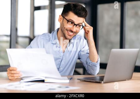 Happy successful confident young caucasian businessman, manager or freelancer wearing glasses sitting at table in office enjoying profit, holding business documents, looking at laptop screen, smiling Stock Photo